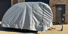 A Little Guy Micro Max trailer is parked on concrete outside a brick building, fully wrapped in a PahaQue Wilderness Little Guy Micro Max Cover, made from durable MagnaTuff fabric with only the hitch and wheels partially visible.