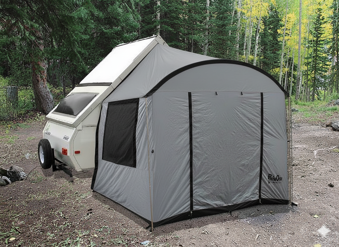 A white Aliner trailer with a Paha Que Custom A-Frame Trailer Side Tent for Aliner Chalet Rockwood Jayco 12 and 15 Foot Box Sizes is set up in a forest clearing, surrounded by trees and sunlight.