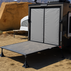A black and gray camper with its rear door open as a ramp is parked on sandy ground near large rocks. It features a Paha Que Custom Toy Hauler Mesh Door/Solid Room Divider. A white tent and folding table are in the background.