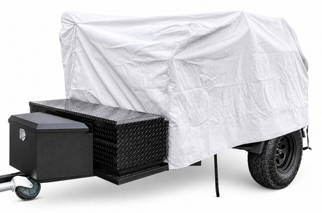 A utility trailer with large black tires is partly protected by the PahaQue Wilderness Vintage Trailer Works XTR Cover, offering heavy-duty weather protection. The front section includes a black lockable box and diamond plate stacked metal toolboxes.