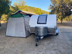 A gray teardrop camper with a Paha Que Custom 5x7 Trailer Side Tent Screen Room is parked by a large tent at a campsite surrounded by trees and a covered pavilion, with camping gear under the sunny, clear sky.