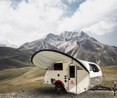 A compact white teardrop camper with a PahaQue Wilderness Little Guy Micro Max Visor awning is parked on grassland before rugged, snow-capped mountains under a partly cloudy sky.