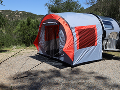 A Paha Que Custom NuCamp TAB 320 Trailer Side Tent for TAB and Boondock Trailers is attached to a camper on gravel, surrounded by trees and hills under a clear blue sky.
