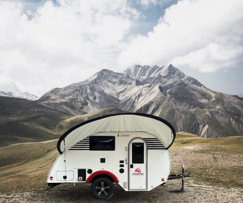 A small white teardrop camper with a PahaQue Wilderness Little Guy Micro Max Visor awning is parked on grass, set before rugged mountains and a partly cloudy sky.