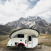 A small white teardrop camper with a PahaQue Wilderness Little Guy Micro Max Visor awning is parked on grass, set before rugged mountains and a partly cloudy sky.