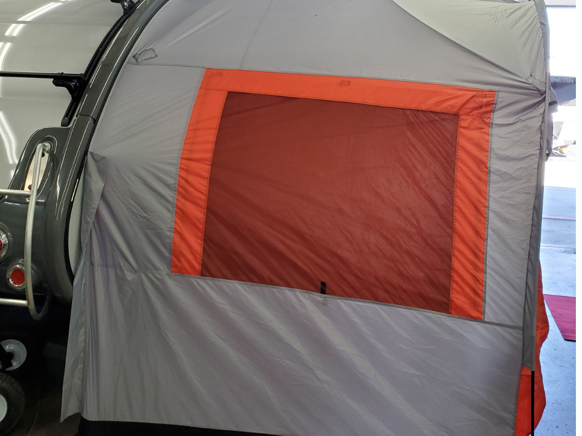 A close-up of the Paha Que Custom NuCamp TAB 320 Trailer Side Tent for TAB and Boondock Trailers, shown in gray and orange with a large rectangular mesh window, attached to a small trailer inside an indoor space.
