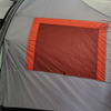 A close-up of the Paha Que Custom NuCamp TAB 320 Trailer Side Tent for TAB and Boondock Trailers, shown in gray and orange with a large rectangular mesh window, attached to a small trailer inside an indoor space.