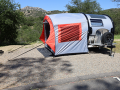 A compact teardrop trailer with a Paha Que Custom NuCamp TAB 320 Trailer Side Tent for TAB and Boondock Trailers is set up on a gravel campsite among trees and hills under a clear sky.