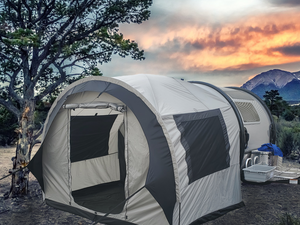 A spacious gray Paha Que Custom NuCamp TAB 320 Trailer Side Tent for TAB and Boondock Trailers is set up outdoors by trees, with camping gear on a table beside it and a mountain sunset in the background—an ideal trailer camping accessory.