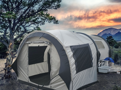 A spacious gray Paha Que Custom NuCamp TAB 320 Trailer Side Tent for TAB and Boondock Trailers is set up outdoors by trees, with camping gear on a table beside it and a mountain sunset in the background—an ideal trailer camping accessory.