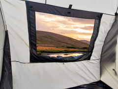 Enjoy the view from inside the Paha Que Custom NuCamp TAB 320 Trailer Side Tent for TAB and Boondock Trailers, with white and black fabric framing a grassy hill, pond, and vibrant sunset—highlighting this unique camping accessory.