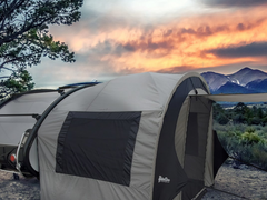A Paha Que Custom NuCamp TAB 320 Trailer Side Tent for TAB and Boondock Trailers stands next to a trailer under a vivid orange sunset, surrounded by shrubs, trees, and distant mountains.