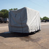 A large vehicle protected by a Paha Que Custom Hiker Trailer Covers Custom Fit Outdoor Protection is parked on an asphalt lot, with other vehicles, trees, and power lines visible in the background under a clear blue sky.
