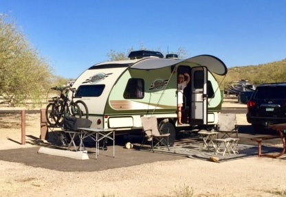 A campsite features a compact trailer with a Paha Que Custom R Pod Trailer Visor. Two bikes are mounted in front, lawn chairs and a table sit outside, and a person stands in the doorway. A car is parked close by.