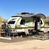 A campsite features a compact trailer with a Paha Que Custom R Pod Trailer Visor. Two bikes are mounted in front, lawn chairs and a table sit outside, and a person stands in the doorway. A car is parked close by.