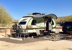 A campsite features a compact trailer with a Paha Que Custom R Pod Trailer Visor. Two bikes are mounted in front, lawn chairs and a table sit outside, and a person stands in the doorway. A car is parked close by.