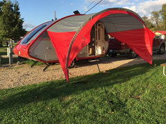 The NuCamp TAB 320 Trailer Awning by PahaQue (Paha Que Custom) is set up in red and gray on grass and gravel, providing shade. A pickup truck is parked behind the camper, with trees and a blue sky in the background.