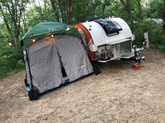 A Paha Que Custom 5x7 Trailer Side Tent Screen Room is set up next to a small white teardrop camper in the woods, with string lights above and camping gear scattered among lush trees at the campsite.