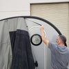 A man in a gray shirt sets up a Paha Que Custom NuCamp TAB 320 Trailer Side Tent for TAB and Boondock Trailers, attaching it to a small trailer with a round window in front of a closed garage door.