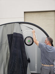 A man in a gray shirt sets up a Paha Que Custom NuCamp TAB 320 Trailer Side Tent for TAB and Boondock Trailers, attaching it to a small trailer with a round window in front of a closed garage door.