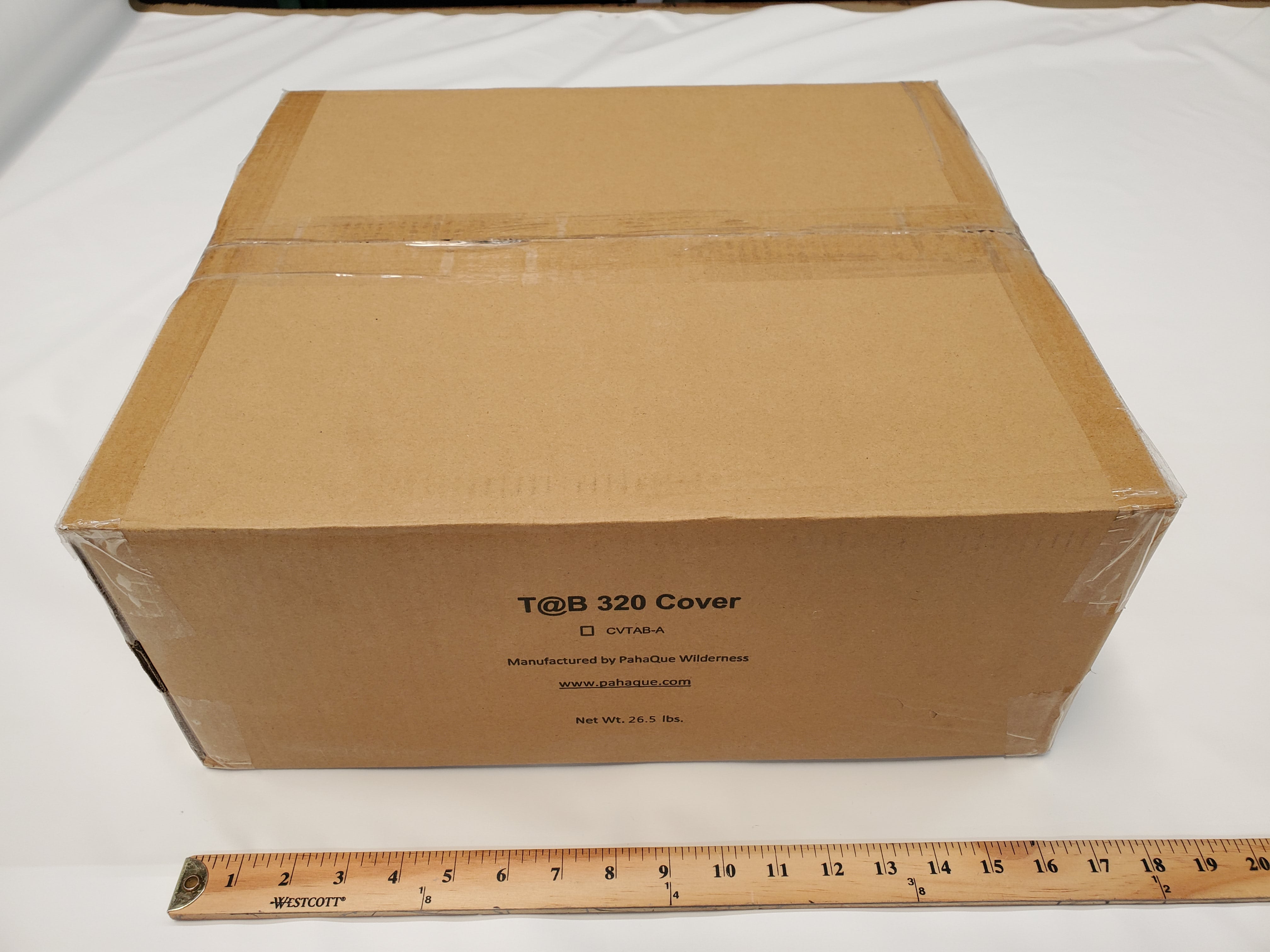 A sealed cardboard box labeled NuCamp TAB 360 CS (Clamshell) Trailer Cover by PahaQue sits on a white surface beside a wooden ruler. This Paha Que Custom product features heavy-duty, weatherproof protection and comes in a box about 24 inches long.
