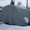 A gray camper is protected by a Paha Que Custom NuCamp TAB 360 CS Clamshell Trailer Cover, offering heavy-duty weatherproof protection. The MagnaTuff fabric cover is topped with snow, with snowy trees and a road seen in the background.