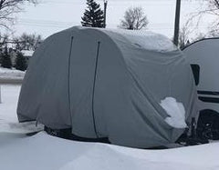A gray camper is protected by a Paha Que Custom NuCamp TAB 360 CS Clamshell Trailer Cover, offering heavy-duty weatherproof protection. The MagnaTuff fabric cover is topped with snow, with snowy trees and a road seen in the background.
