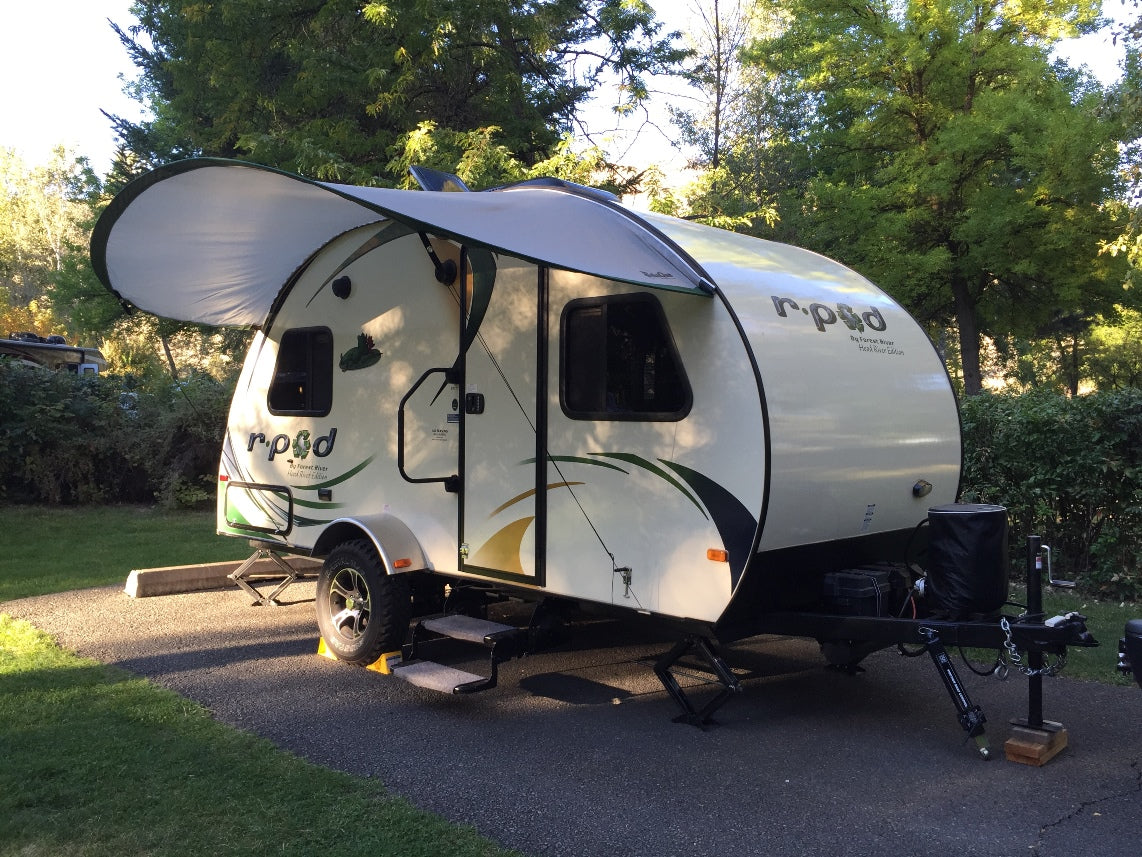 A small white R-Pod trailer with green and yellow accents is parked on pavement in the woods. Out front, a Paha Que Custom R Pod Trailer Visor provides extra shade beneath the awning amid lush trees.