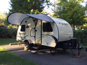 A small white R-Pod trailer with green and yellow accents is parked on pavement in the woods. Out front, a Paha Que Custom R Pod Trailer Visor provides extra shade beneath the awning amid lush trees.