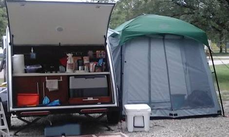 A small camper with its back hatch open shows a compact kitchen setup beside a Paha Que Custom 5x7 Trailer Side Tent Screen Room for Camping and Outdoor Adventures, with camping gear and containers set up on gravel.