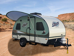 A compact, modern travel trailer equipped with a Paha Que Custom R Pod Trailer Visor is parked on a sandy dirt path in the desert, its extended awning offering shade among rocky hills and scattered vegetation.